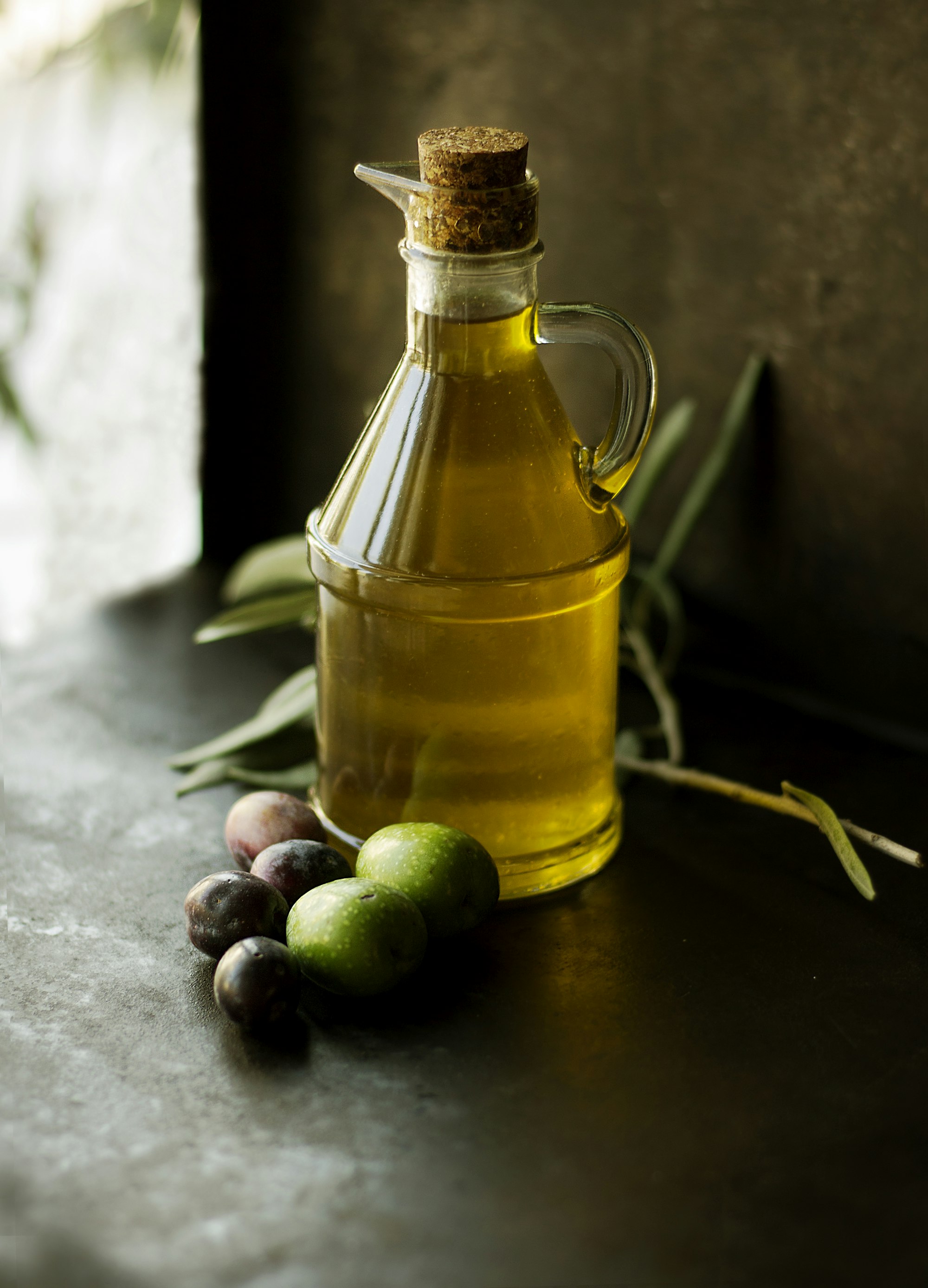 Glass bottle of amber liquid set on a dark countertop beside fresh olives and leaves — a quiet, dimly lit pantry still life.
