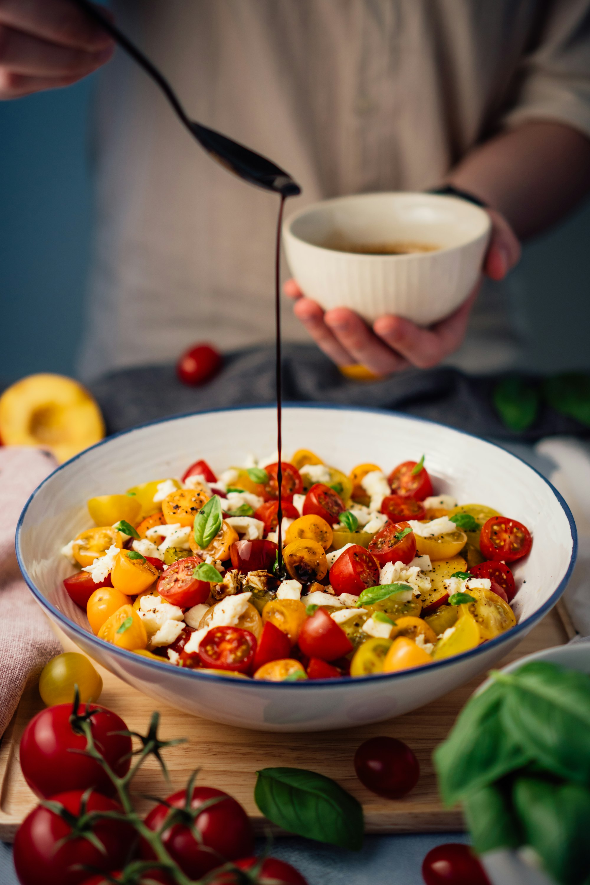 A heritage tomato and mozzarella salad finished with a slow stream of dark vinegar from a small bowl — fresh basil and ripe tomatoes alongside.